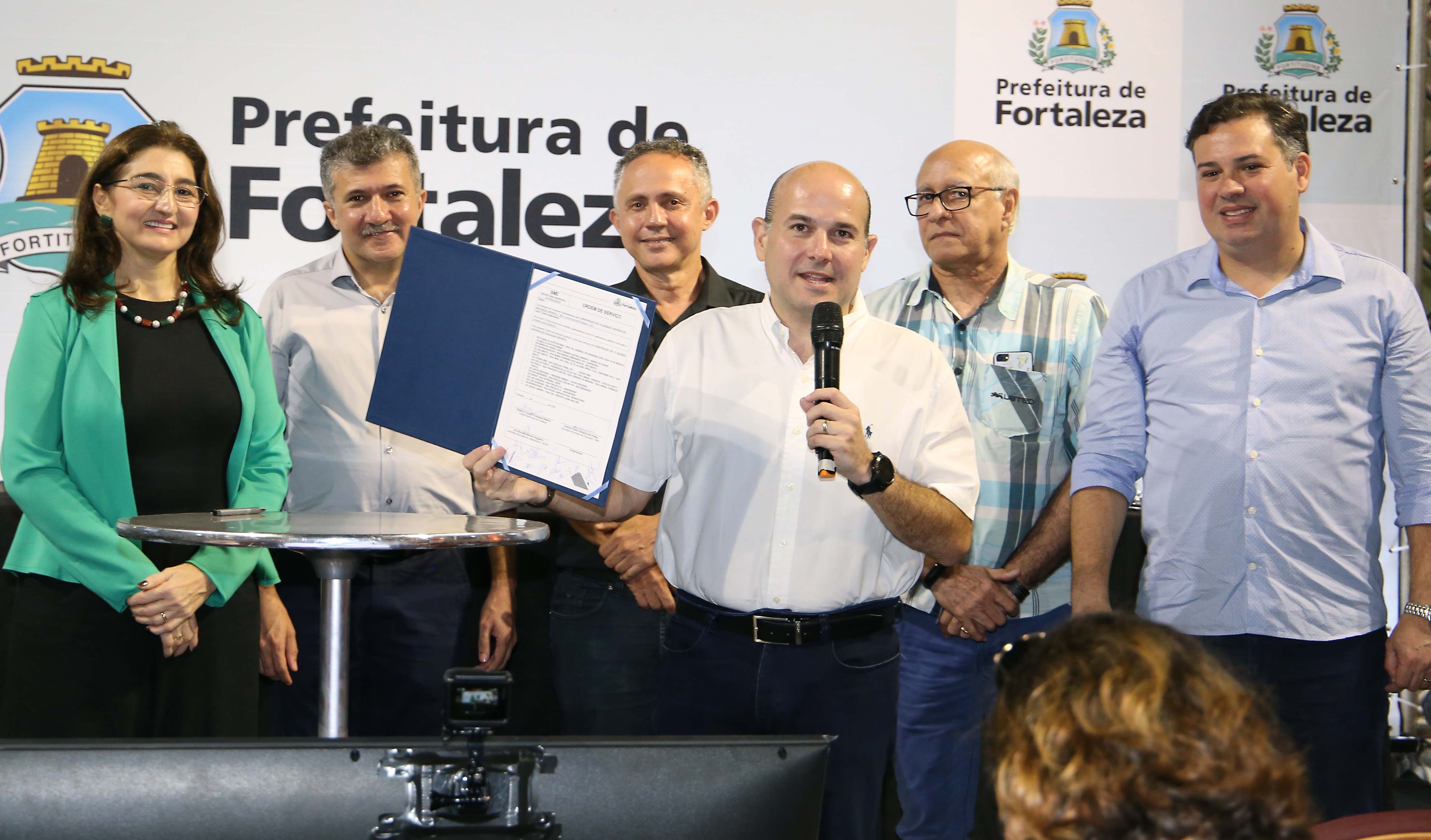 prefeito em pé falando ao microfone mostrando documento e cinco pessoas atrás em pé